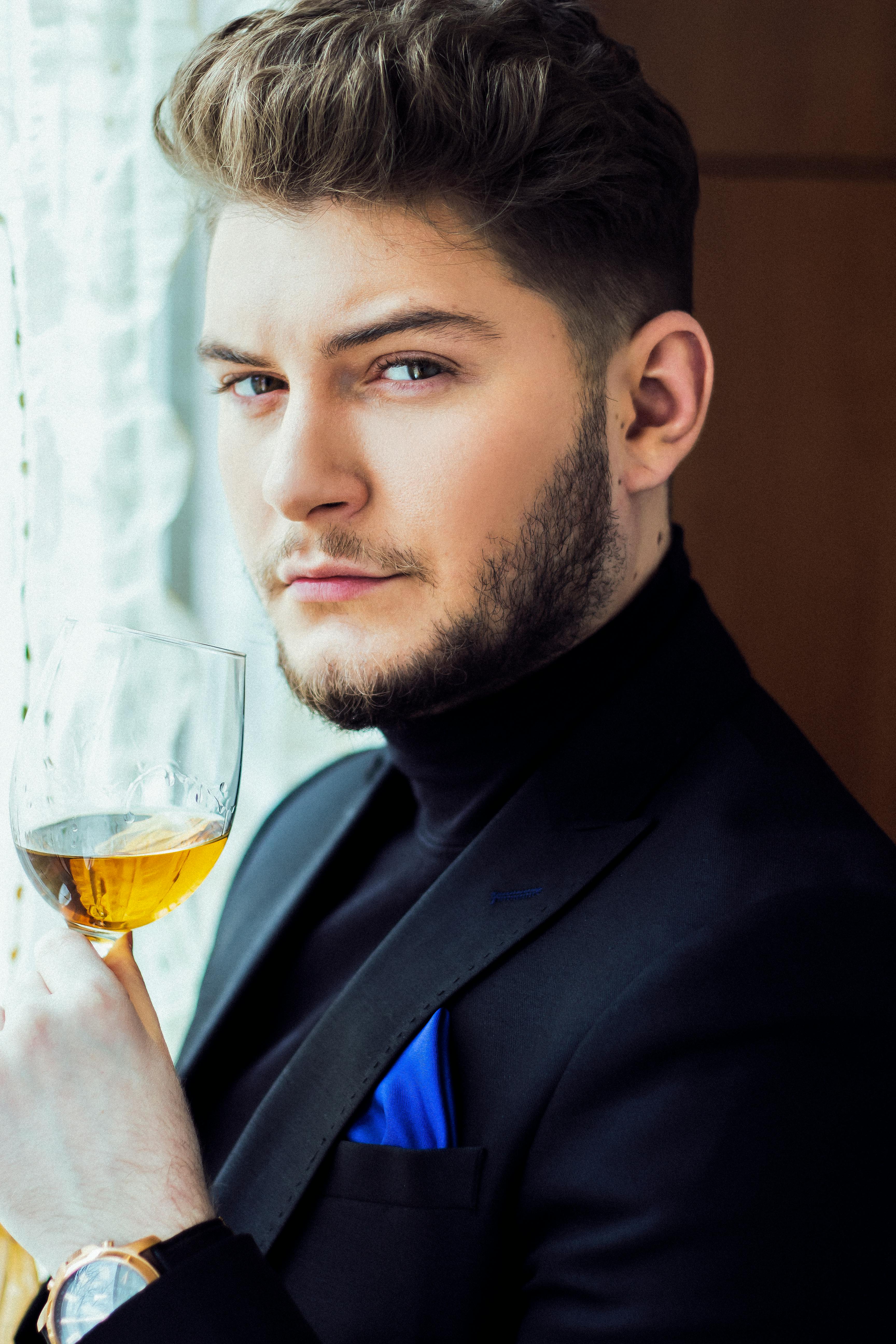 Serios bearded male in elegant suit standing near window with glass of champagne and looking at camera while resting during party