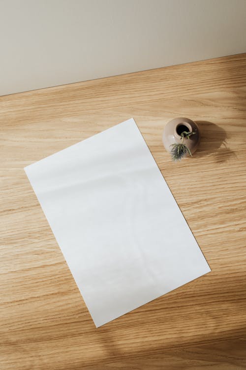 From above composition of empty white paper sheet placed near beige creative ceramic vase with artificial flower on wooden desk against white wall in daytime