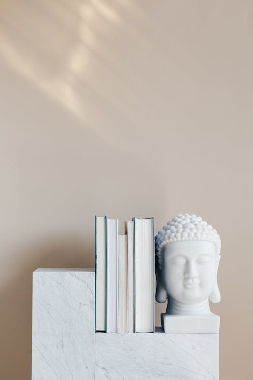 Set of various books composed with white Buddha head statuette on marble stand against beige wall