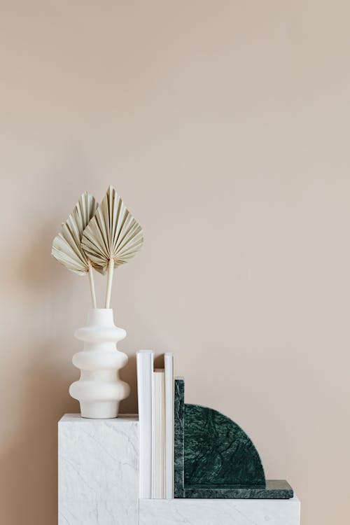 Minimalistic interior design with original vase with decorative golden leaves and set of magazines with green marble bookend placed on white shelf