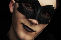 Handsome young male with makeup and black Colombina mask on face looking away on black background