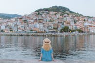 一名身穿蓝色连衣裙的女子坐在码头上，凝视着一座风景如画的沿海小镇