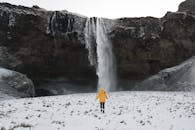 Person in Red Jacket Standing on White Snow Covered Ground Near Waterfalls