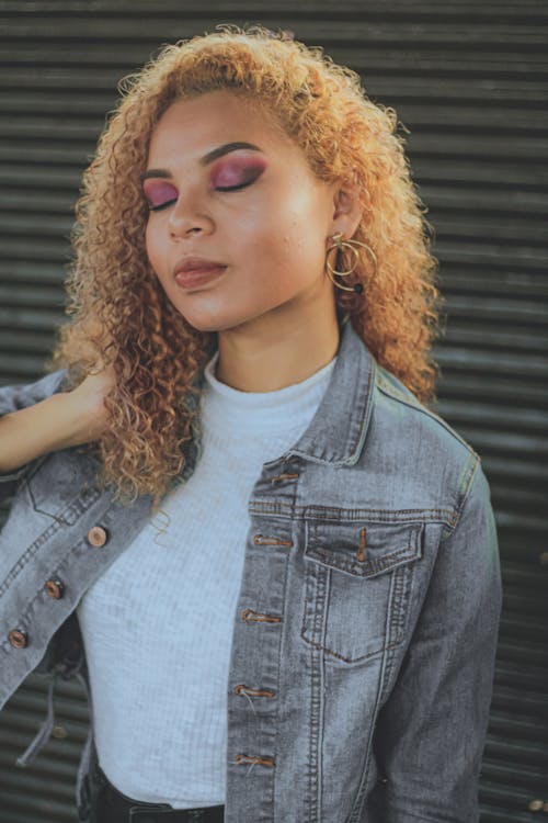Free Unemotional young beautiful ethnic female with golden curly hair and bright makeup in denim coat touching hair while standing next to striped gray wall Stock Photo