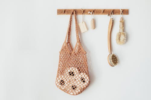 Composition with assorted wooden body brushes of different sizes with organic soap and string bag filled with toilet paper rolls hanging on wooden hanger on white wall in bathroom
