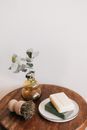 From above composition with assorted organic soaps on white ceramic holder and shaving brush with wooden handle near yellow transparent vase with plant on wooden round table against white wall
