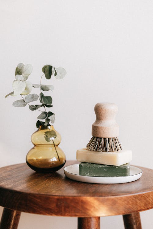 Free Composition of shaving brush placed on organic soaps on ceramic white holder near yellow transparent vase with plant on wooden round table against white wall Stock Photo