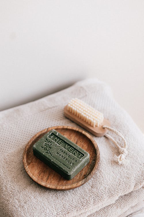 From above composition with natural green soap on eco friendly wooden holder and sustainable brush lying on folded clean soft fresh gray towels against white wall