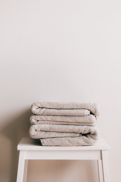 Free Minimalist composition of white simple stool with clean fresh folded gray towels against beige wall in bathroom Stock Photo