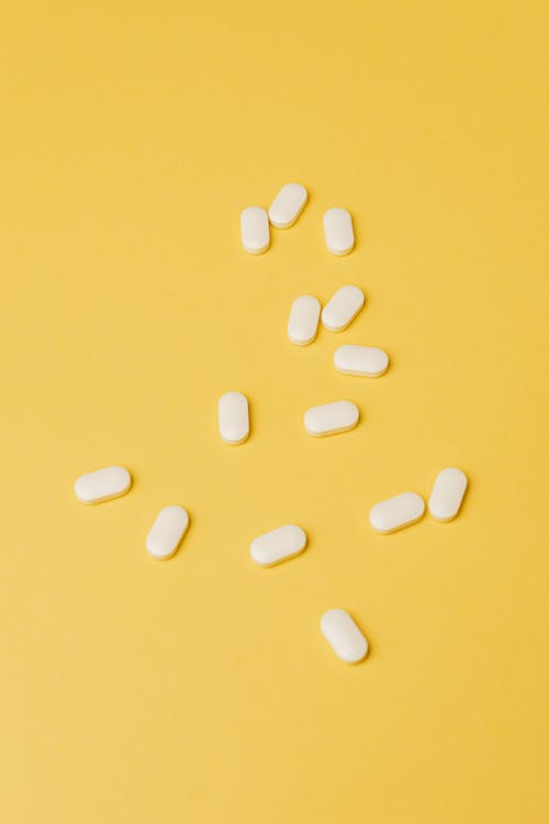 From above heap of elliptical medical white pills placed in disorder on vivid yellow background surface