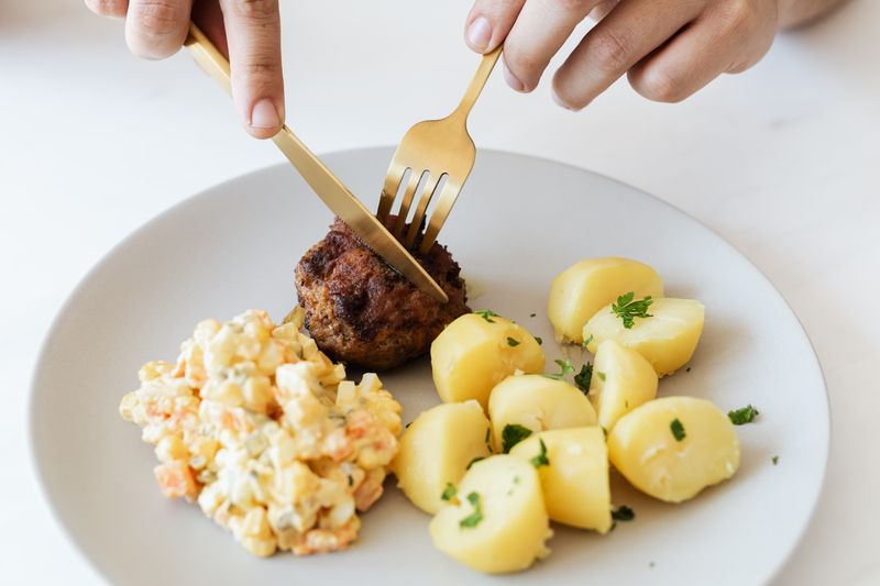 From above of crop anonymous person cutting with cutleries fried juicy meat cutlet served on white plate with tasty boiled potatoes and Russian salad