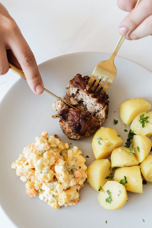 From above of crop anonymous person eating yummy fried beef cutlet served with tasty boiled potatoes and traditional Russian salad with meat potato and green peas