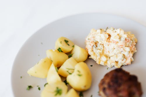 From above of plate with juicy fried meat cutlet served with delicious Russian meat salad and boiled potatoes topped with parsley placed on white table