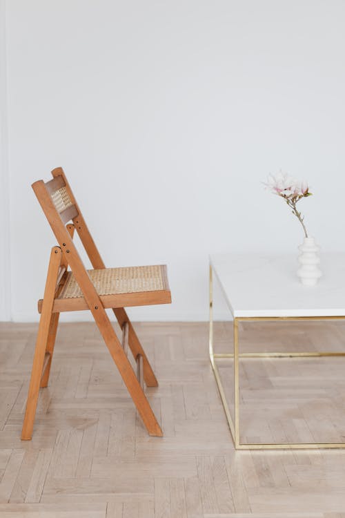 Comfortable wooden net chair located near white top table with vase and pink flower placed on light beige parquet against plain white wall