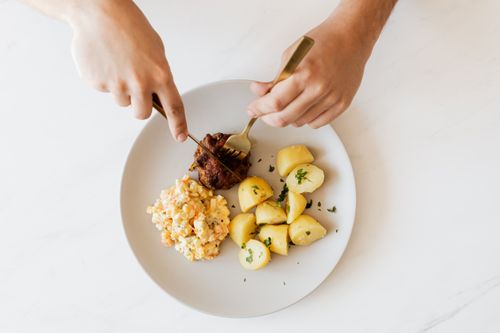 Free Top view of crop unrecognizable male with cutleries slicing delicious beef cutlet garnished with boiled potatoes and parsley and portion of Russian meat salad on plate placed on white table Stock Photo