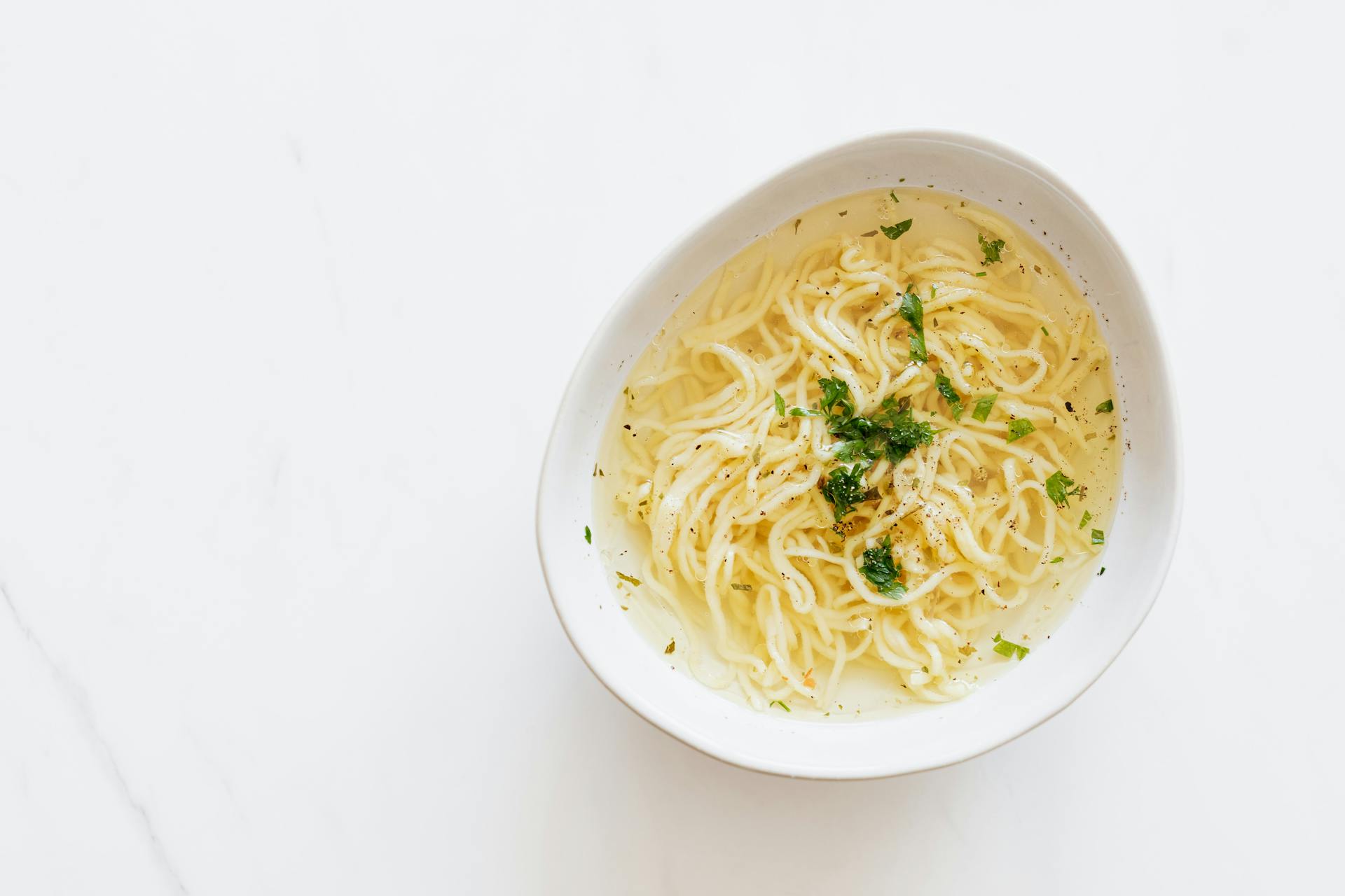 Top view of white bowl with yummy homemade noodles cooked in fresh delicious chicken broth and topped with green aromatic parsley placed on white marble table