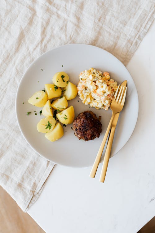 Overhead view of appetizing boiled potatoes sprinkled with greens with yummy rissole and cream vegetable and egg salad served on white plate with gold fork and knife on linen napkin