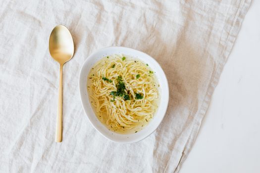 Top view of bowl with yummy noodle soup with greens and golden spoon placed on rumpled linen cloth on marble table