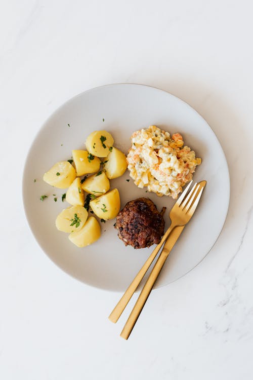 Free Top view of boiled potatoes with vegetable salad with yummy beef cutlet served on white round plate with golden fork and knife on marble table Stock Photo