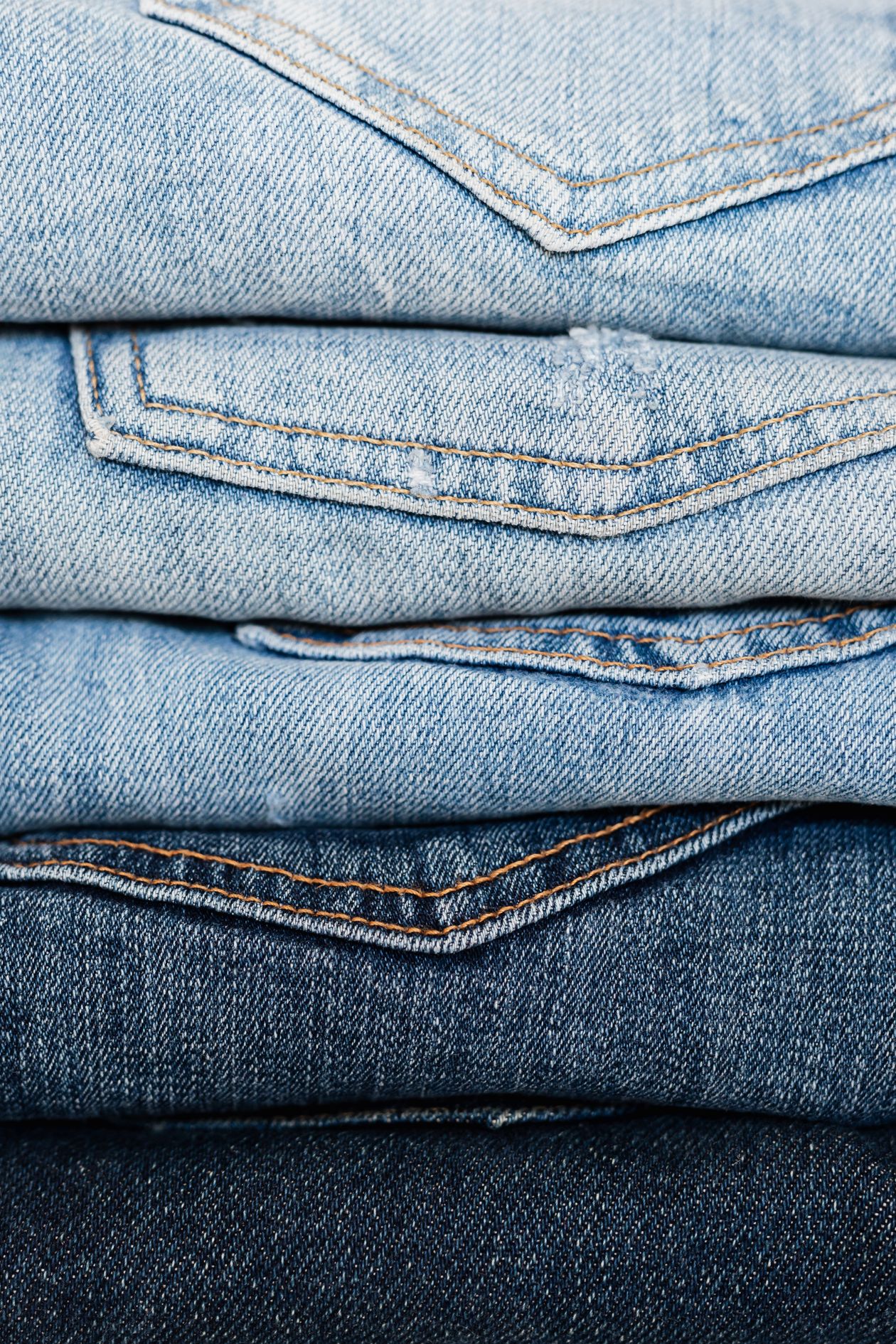Five pairs of folded jeans stacked in a gradient from pale washed blue at the top to raw dark indigo at the bottom, showing the range a wash recipe can move a fabric across.