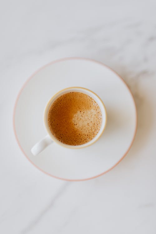 Top view of white cup of coffee with thin froth on white saucer placed on white marble table