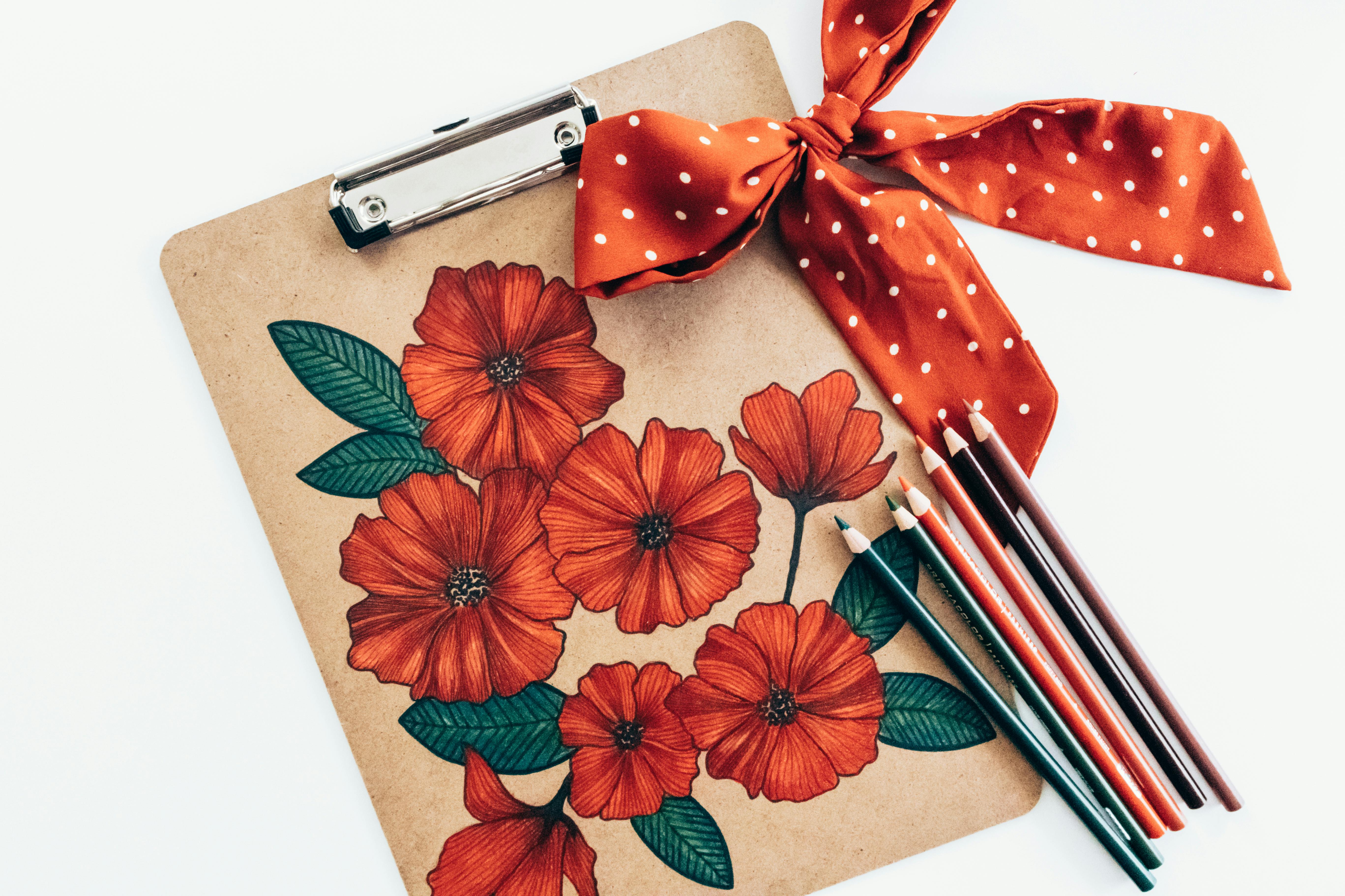 Overhead view of long multi colored pencils and red polka dot bow with ribbon on carton clipboard decorated with picture of red flowers with green leaves on white background