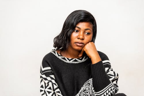 Young upset black female in stylish knitted sweater sitting leaned on hand near wall while looking away