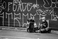 Black and white of anonymous traveler in sterile mask and eyewear sitting on road near ribbed graffiti wall and backpack during COVID 19 pandemic