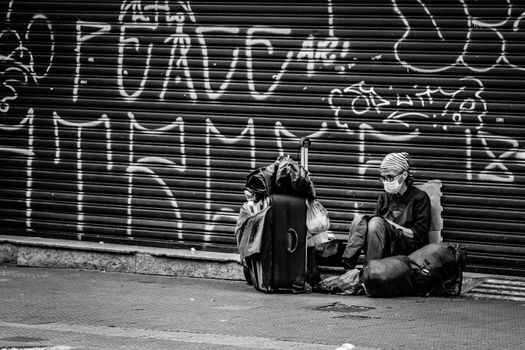Black and white of anonymous traveler in sterile mask and eyewear sitting on road near ribbed graffiti wall and backpack during COVID 19 pandemic