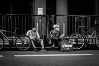 Black and white of anonymous delivery male in face masks sitting near fence and roadway with parked bicycles in town during COVID 19 pandemic