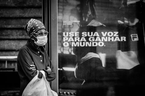 Free Black and white of unrecognizable ethnic female in eyeglasses and sterile mask standing with folded arms near glass wall with inscription during coronavirus pandemic and looking away Stock Photo