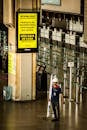 From above of anonymous worker in mask and protective helmet walking with ladder on floor under yellow signboard with information about COVID 19 pandemic