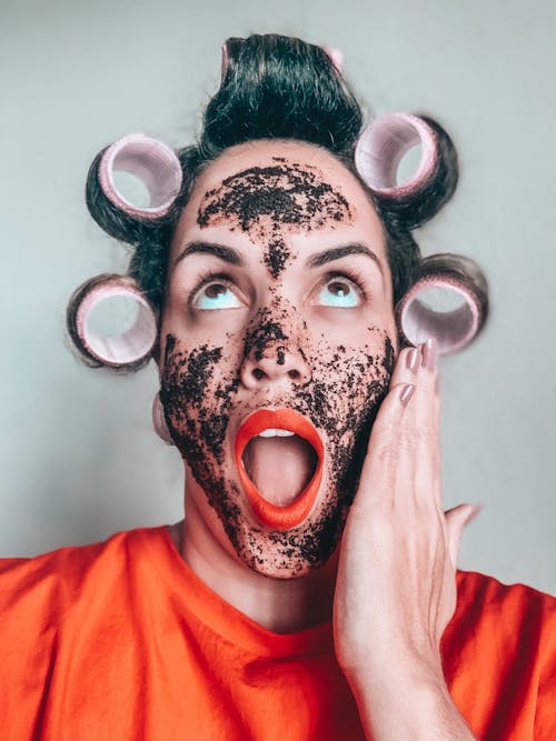 Amazed woman in curlers applying black mask on face