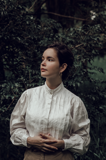 A woman in a white dress shirt stands elegantly in a lush outdoor setting, embodying tranquility and grace.