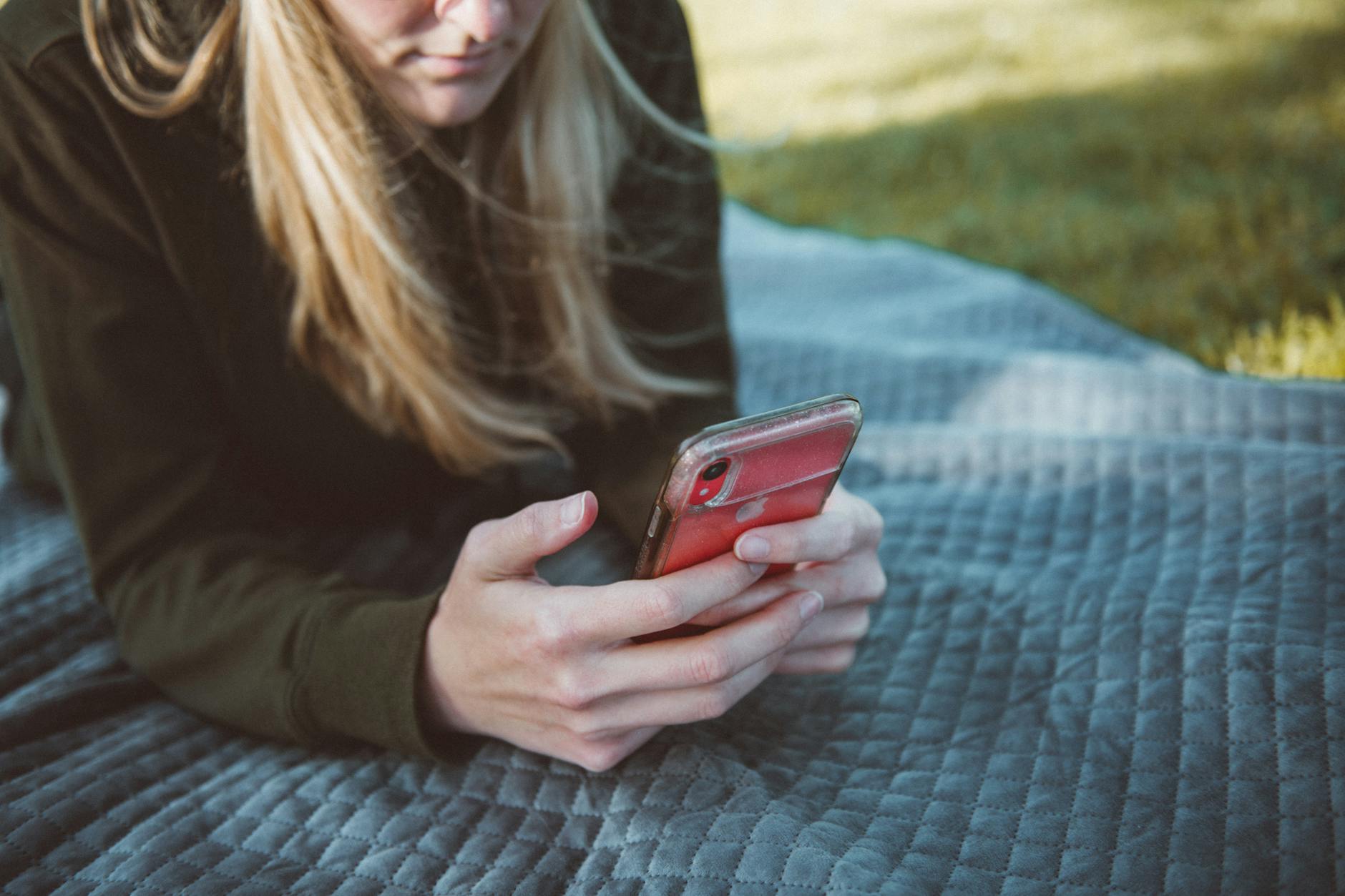 Młoda blondynka leżąca na kocu na świeżym powietrzu, trzymająca w rękach smartfon w czerwonym etui