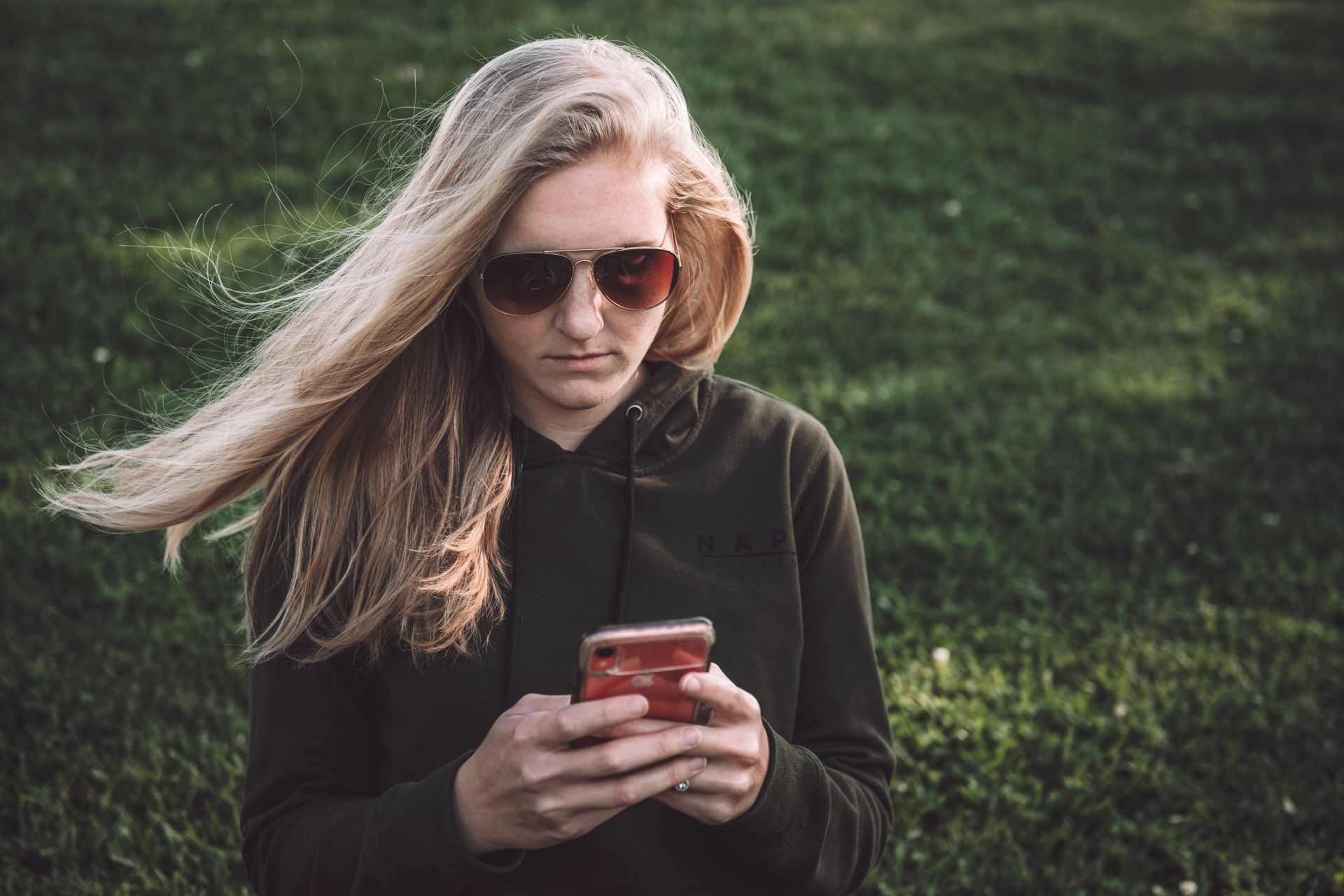 Blonde woman with sunglasses texting on smartphone in a sunny outdoor park.