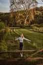 From above of young happy female with reached arms walking on green meadow behind mountain and looking at camera