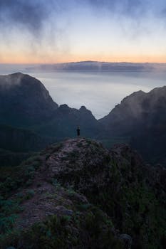 Lone hiker stands on mountain peak, admiring tranquil sunset view.