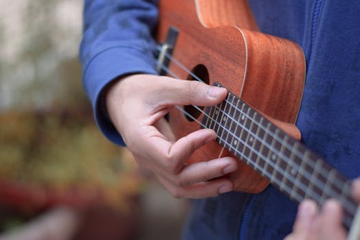 Person in a blue jacket playing a wooden ukulele with focus on hands and strings.