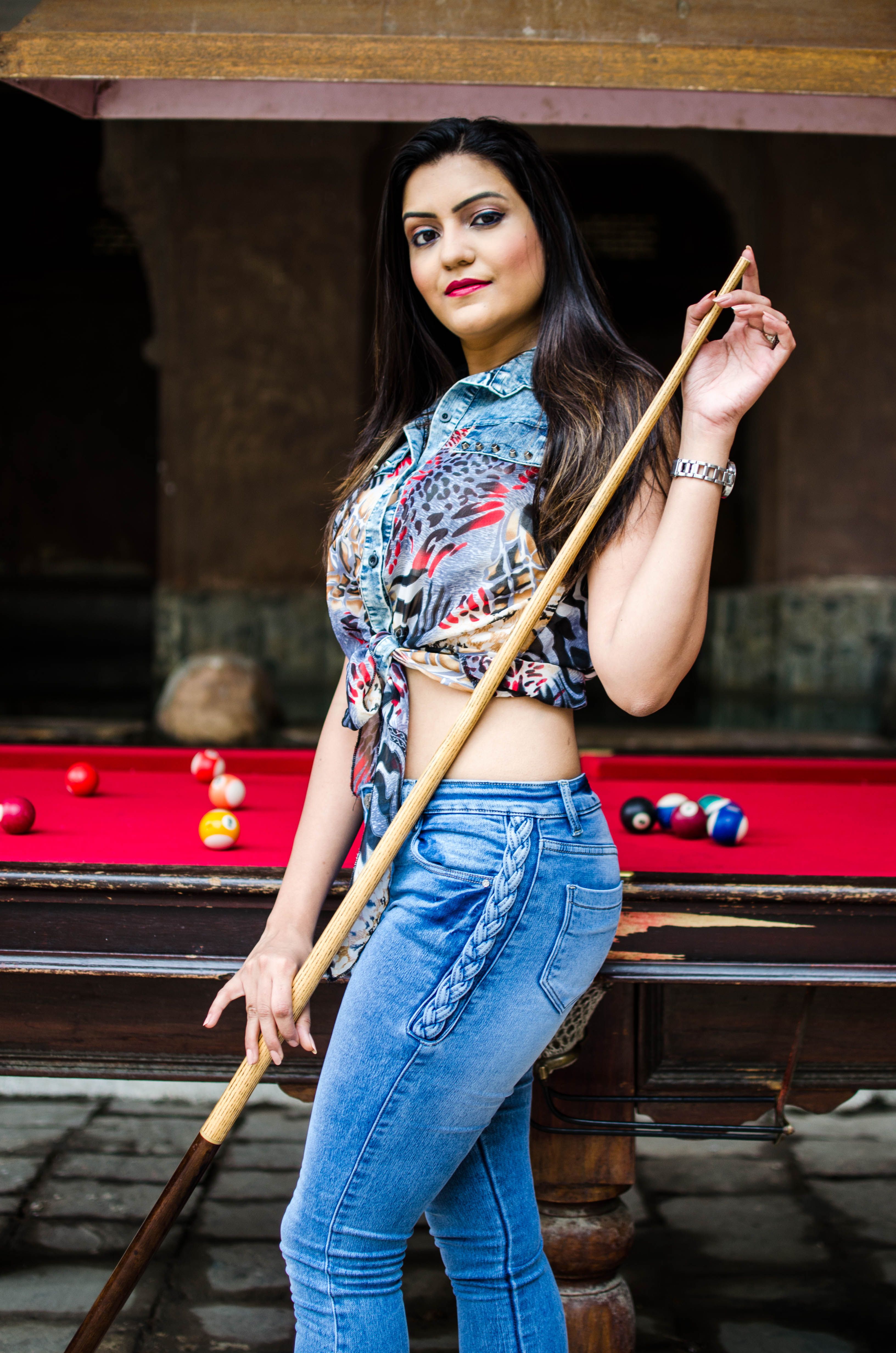 Stylish woman in denim and crop top posing confidently with a billiard cue indoors.