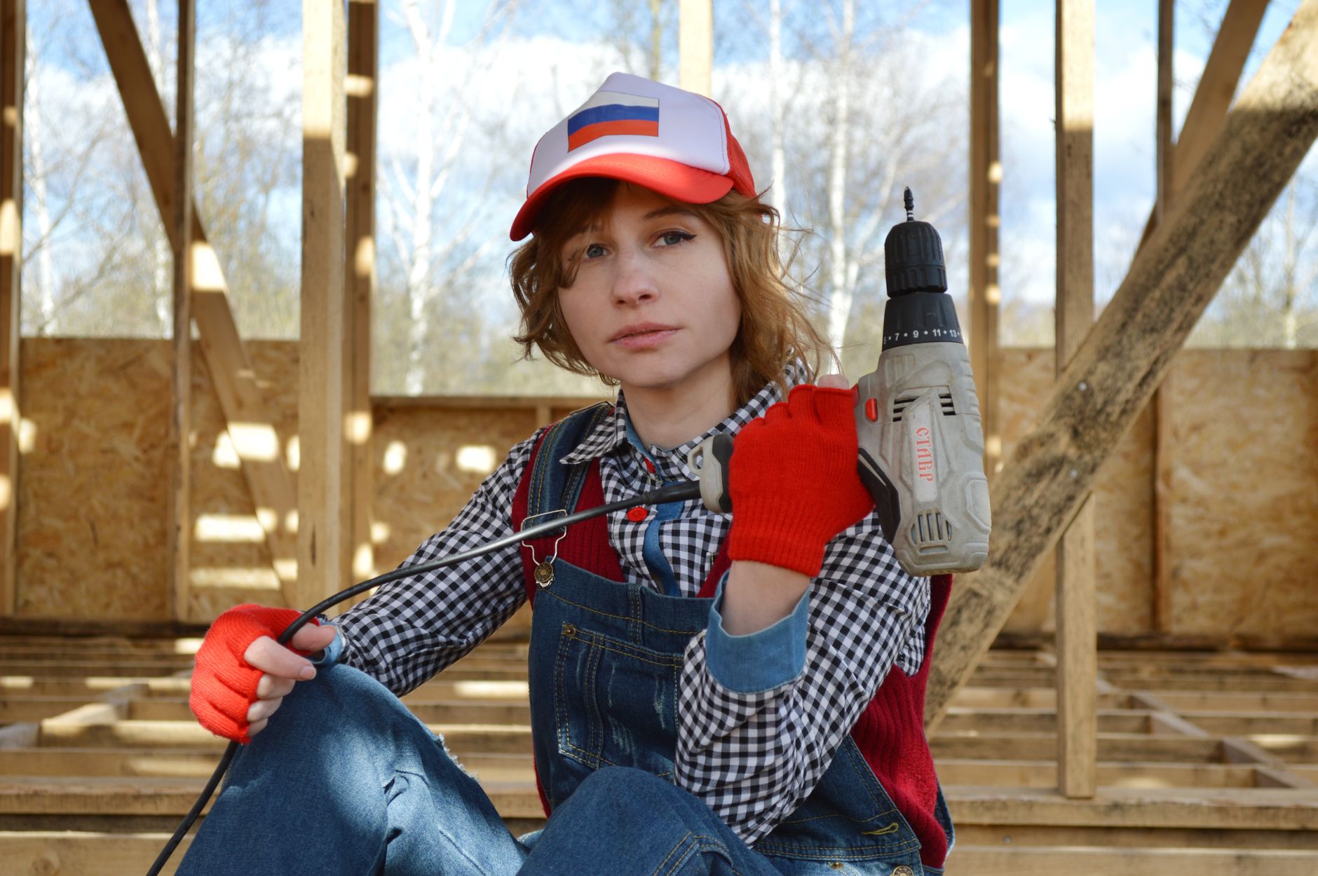 Confident young woman in a checkered shirt and denim, holding a drill on a building site.