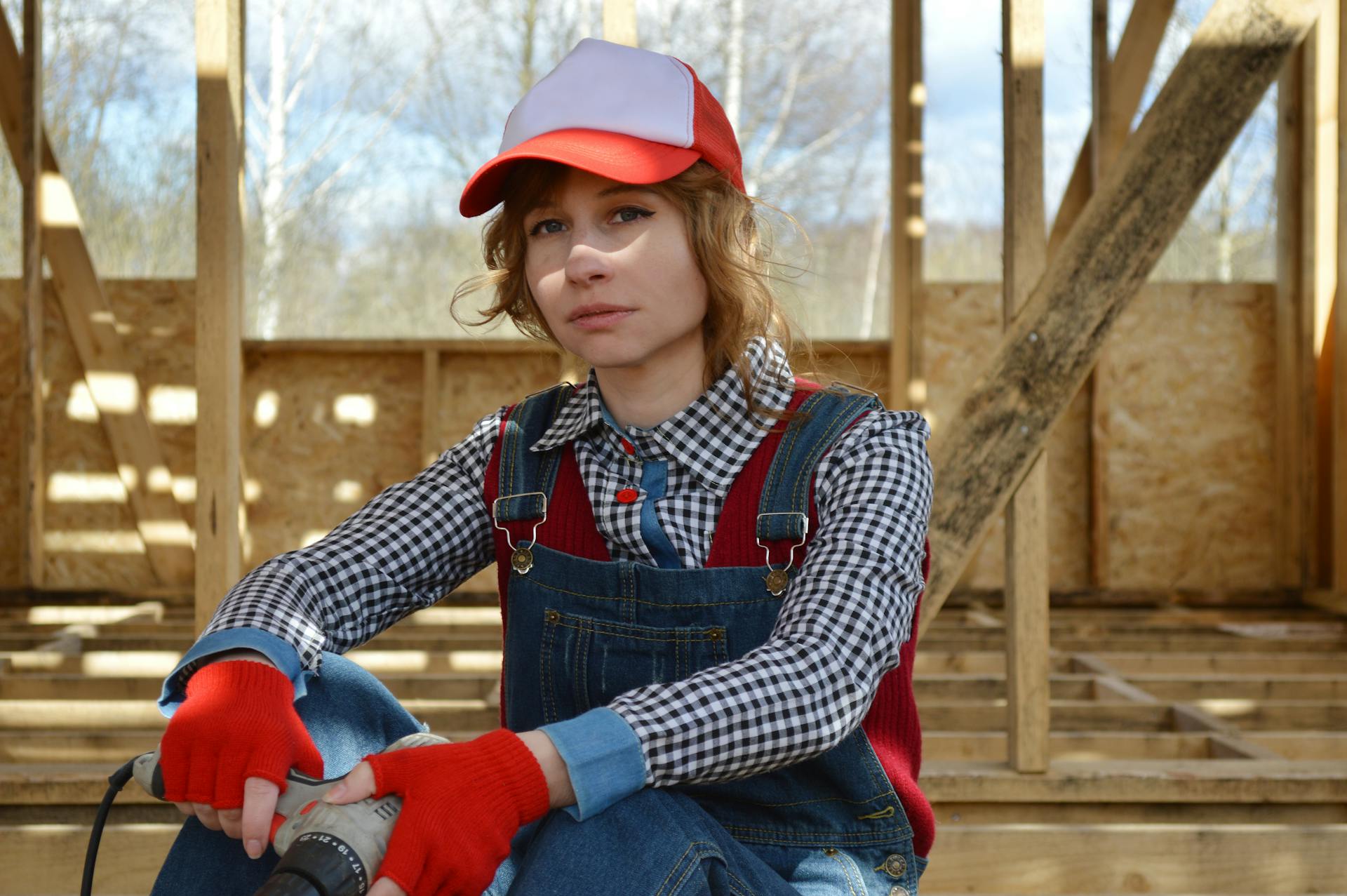 Pondering young female in colorful cap and denim overalls sitting with drill on wooden floor of construction while looking at camera