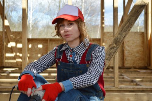 Free Pondering young female in colorful cap and denim overalls sitting with drill on wooden floor of construction while looking at camera Stock Photo