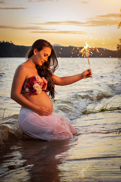 Side view of smiling pregnant lady on coast near wavy ocean with bright Bengal light at sunset