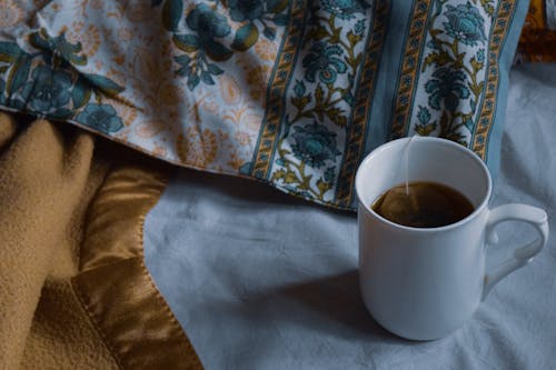From above of white mug of hot drink placed on bed near blanket and pillow