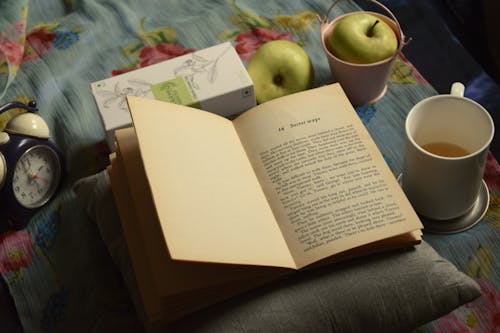 Free High angle composition of interesting book placed on soft pillow near cup of aromatic tea and green apples and alarm clock Stock Photo