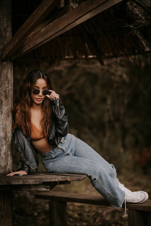 Full body trendy young female in jeans bra and leather jacket resting on abandoned wooden cottage terrace and lowering sunglasses while looking at camera