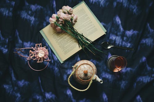 Top view composition of opened book and delicate pink flowers placed on dark blue silk cover near teapot glass of tea and creative candleholder