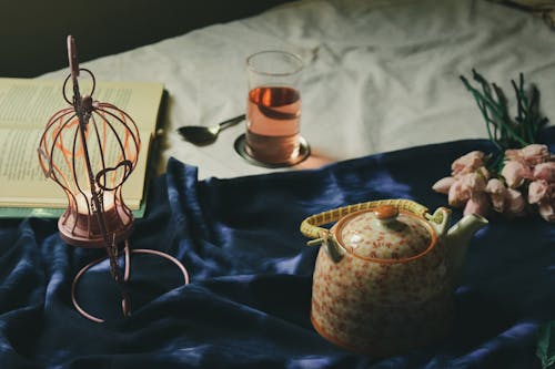Free Arrangement of teapot and creative hanging candleholder with burning tealight candle placed on comfy bed near opened book and glass with drink Stock Photo