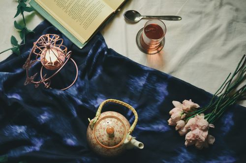 Free Top view composition of ceramic teapot and glass of fresh tea served on cozy bed near delicate pink flower burning tealight candle and interesting book Stock Photo